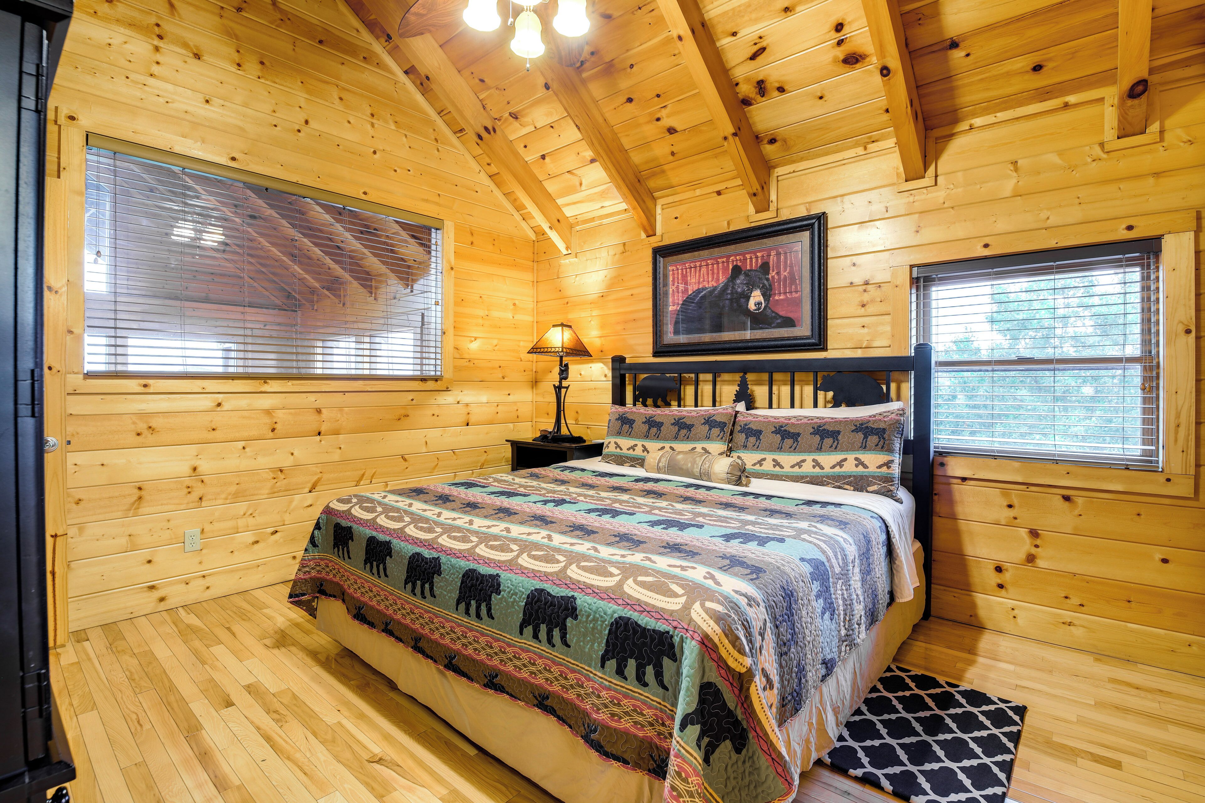 A wooden cabin bedroom with a large bed, a framed bear picture, and a window with blinds.