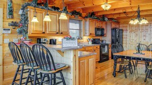 A cozy cabin-style kitchen with wooden cabinets, a central island with bar stools, and festive decorations. - Hot Tub + Games! Mtn-view Cabin Near Pigeon Forge (Sevierville)