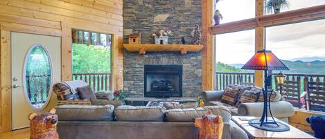 A cozy cabin interior with a stone fireplace, wooden walls, a large window, and a comfortable seating area.