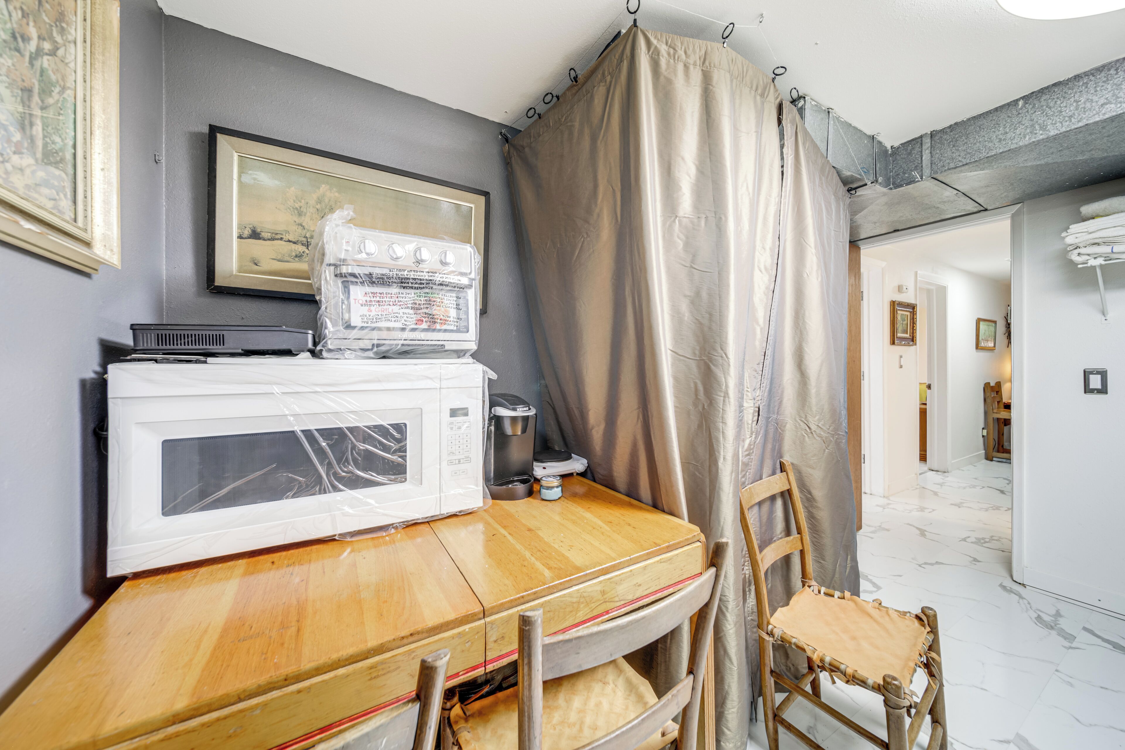 A room with a wooden table, a white microwave, a chair, and a beige curtain.