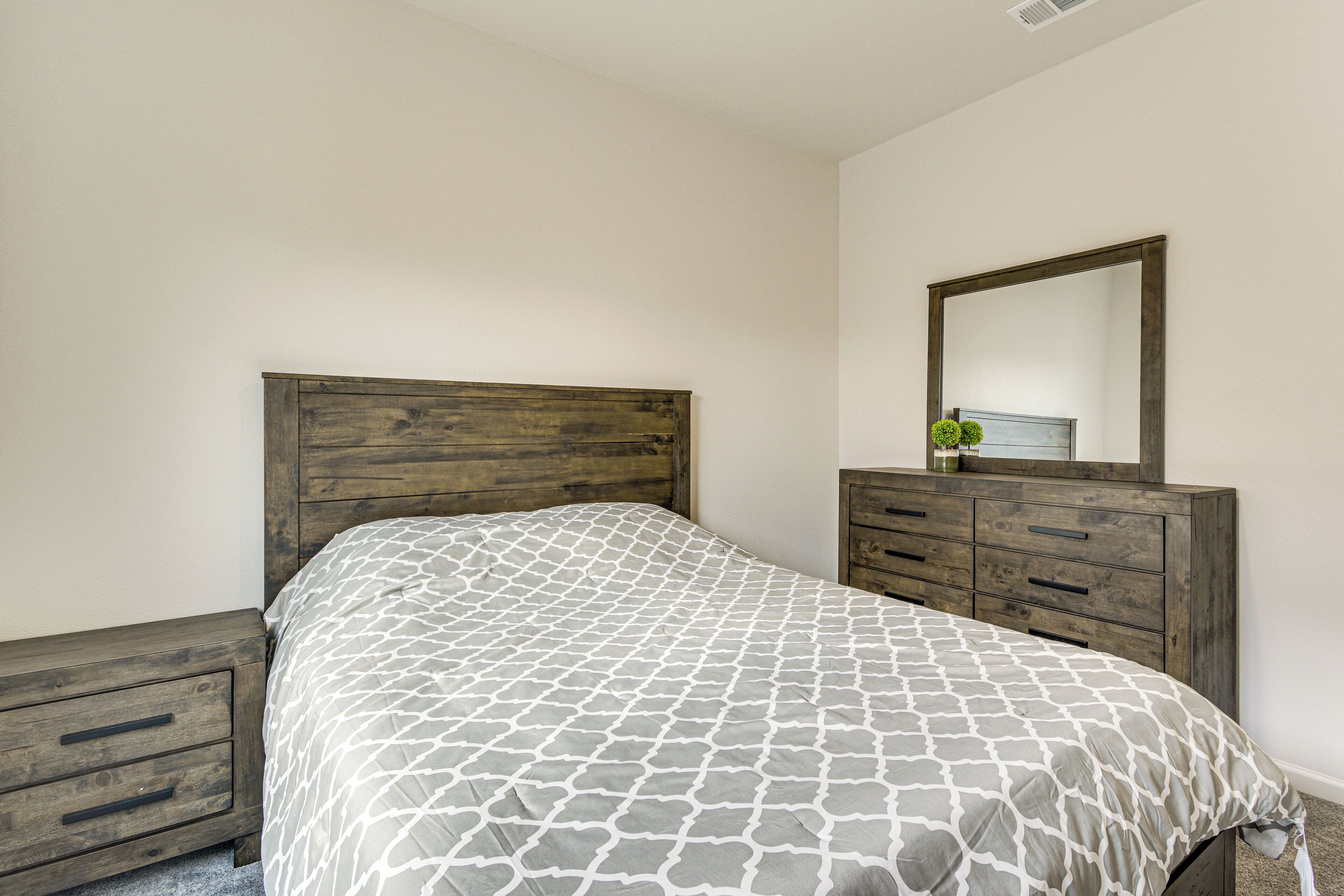 A bedroom with a wooden headboard, a bed with a patterned bedspread, a dresser with drawers, and a mirror on the dresser.