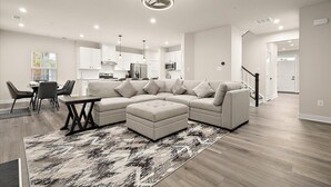 A modern living room with a sectional sofa, a dining area with chairs, a kitchen in the background, and a staircase leading upstairs.