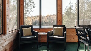 A cozy cabin interior with two wicker chairs, a small table, and large windows offering a view of a forest and lake.