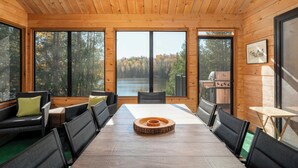 A wooden cabin with a dining table and chairs, large windows with a forest view, and a grill outside.