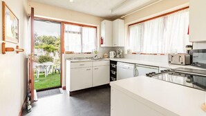 A compact kitchen with white cabinets, a dishwasher, a stove, and a sink. There is a small dining area with a table and chairs visible through an open door. - Host Stay Cransley (Great Yarmouth)