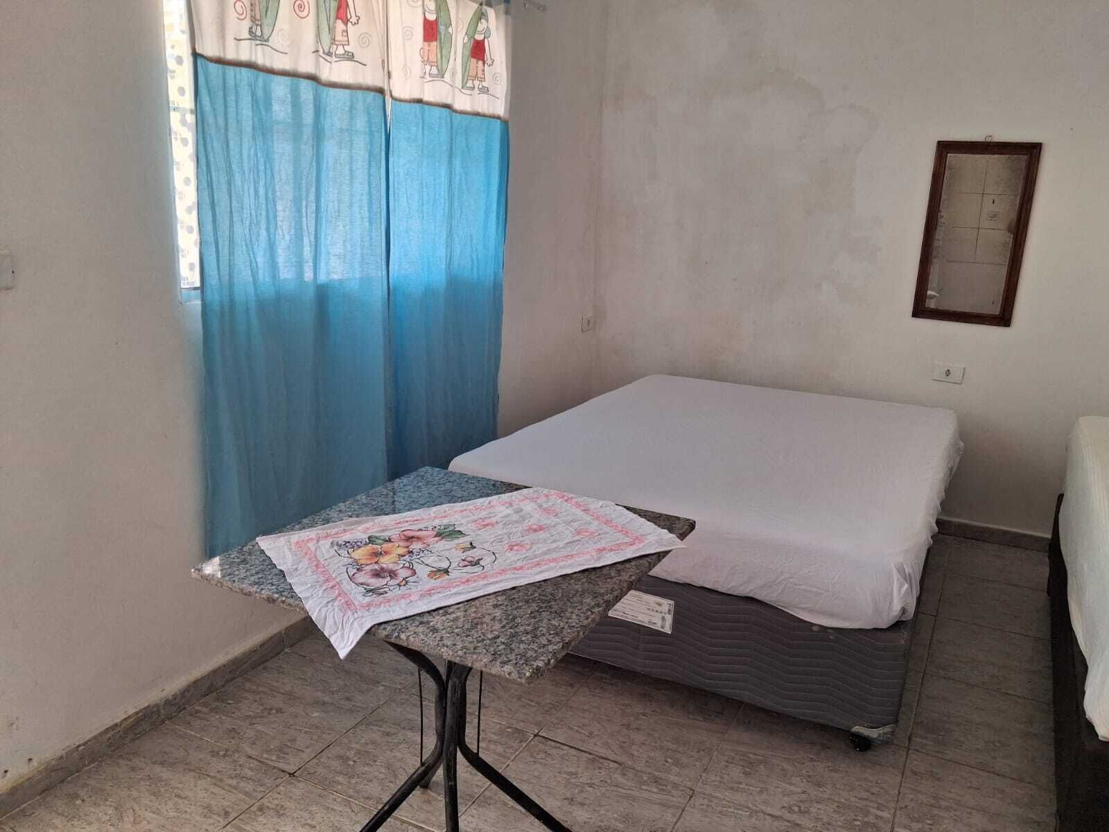 A small, simple room with a bed, a table with a floral tablecloth, and a window with blue curtains.