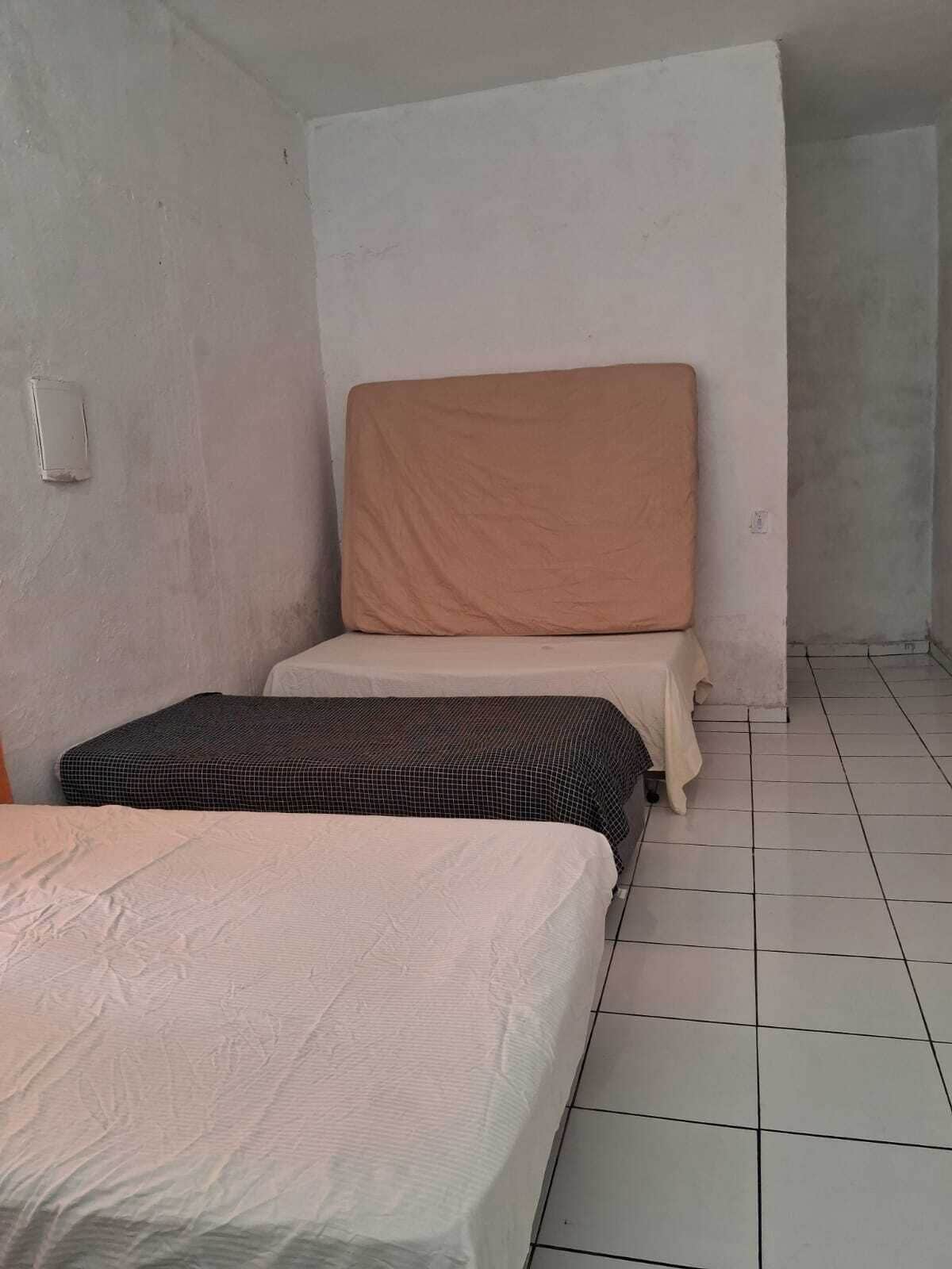 Una habitación pequeña y sencilla con dos camas, una con una colcha estampada y la otra con una lisa. Las paredes son lisas y el suelo, de baldosas.