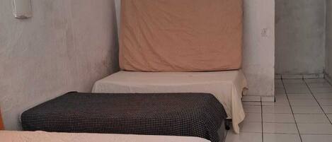 A small, simple room with two beds, one with a patterned bedspread and the other with a plain one. The room has plain walls and a tiled floor.