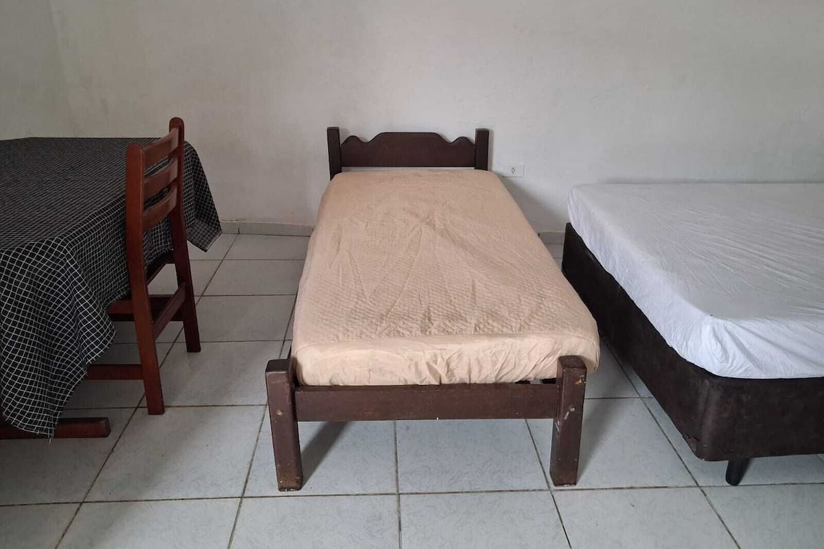 Ein kleines Zimmer mit einem Einzelbett, einem hölzernen Bettgestell, einem passenden Holzstuhl und einem kleinen Tisch mit kariertem Tischläufer.