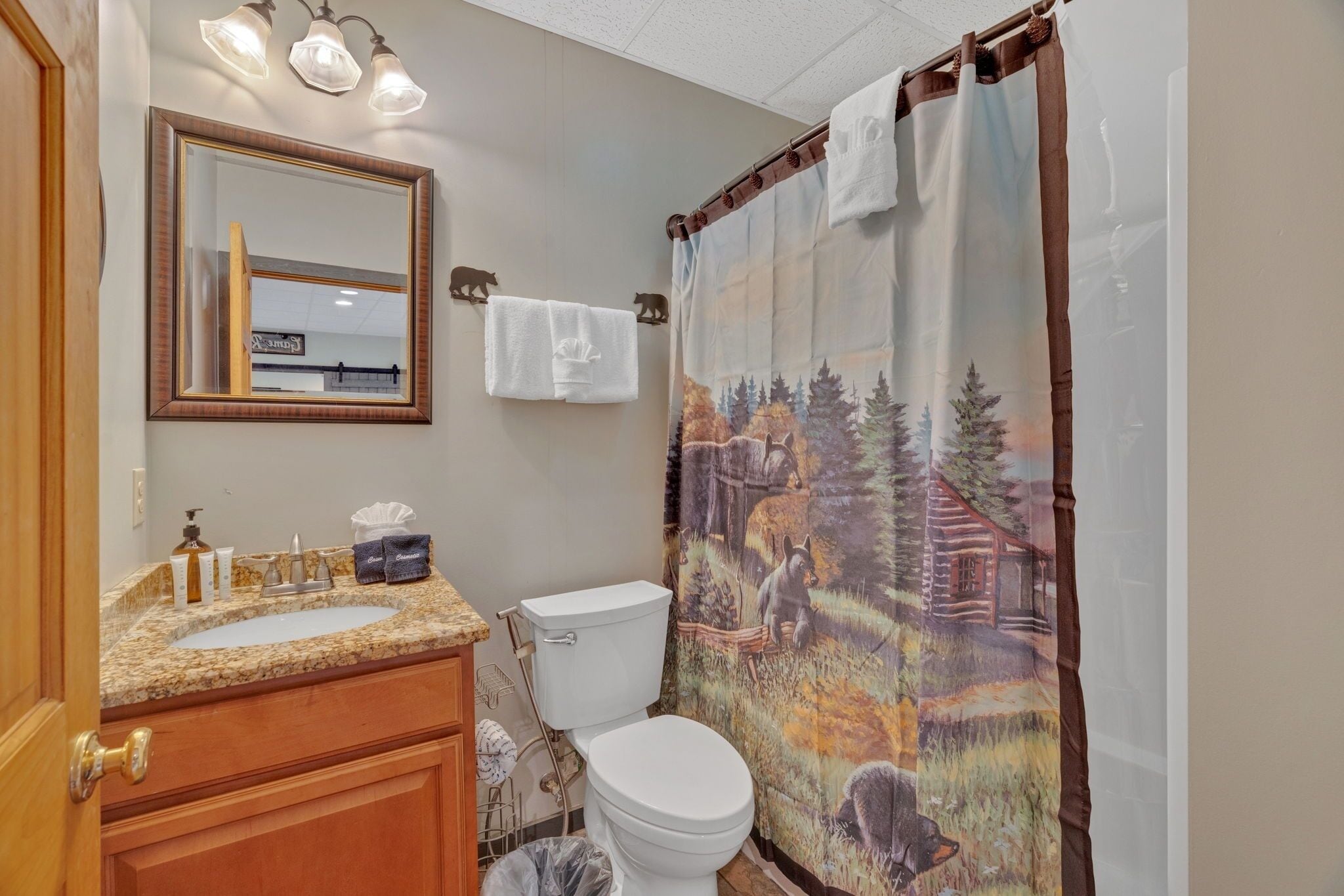 A bathroom with a sink, mirror, toilet, and shower curtain with a forest scene.
