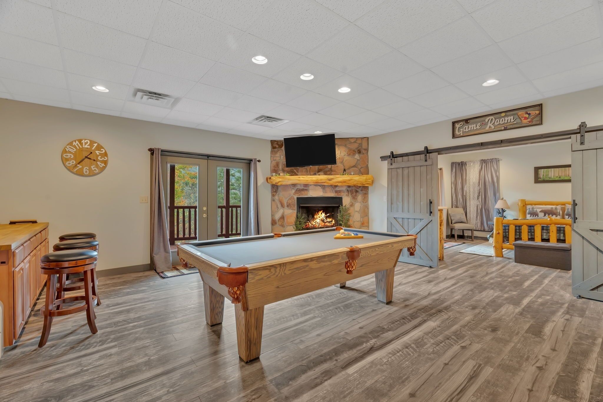 A pool table in a room with a stone fireplace, wooden flooring, and a clock on the wall.