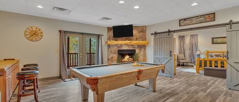 A pool table in a room with a stone fireplace, wooden flooring, and a clock on the wall.