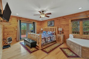 A wooden cabin bedroom with a bed, a ceiling fan, a TV, a window, a bathroom, and a view of the outdoors. - Walden's Ridge Cabin 4 Bedroom Cabin (Sevierville)