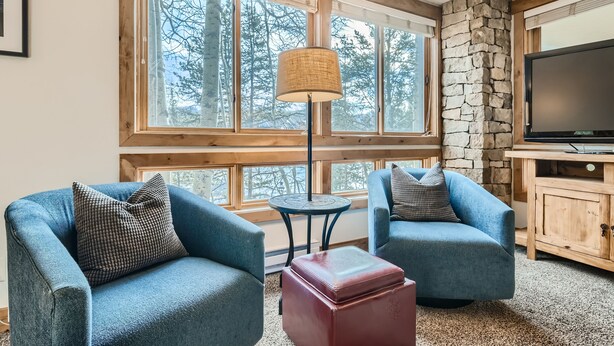 A cozy room with two blue armchairs, a small table, a red ottoman, a stone wall, a large window with a view of trees, and a television on a wooden cabinet.