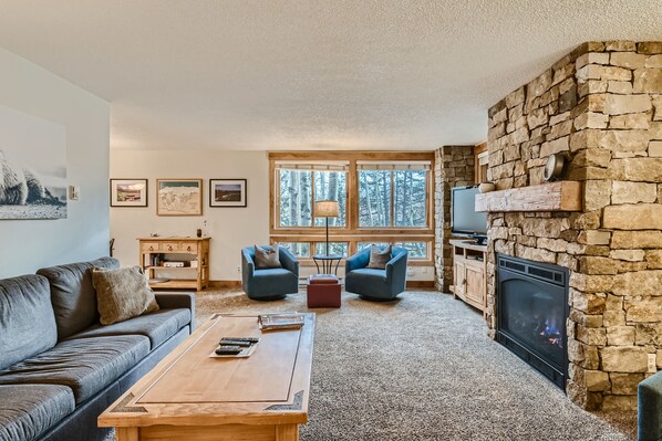 A cozy living room with a stone fireplace, a sofa, two armchairs, a coffee table, and a TV. - Ski Hill 23 2 Bedroom Condo (Breckenridge)
