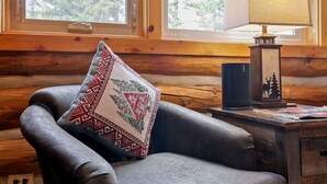 Interior de una cabaña de troncos con un sillón de cuero, un cojín decorativo estampado, una lámpara y una mesita con un objeto decorativo.