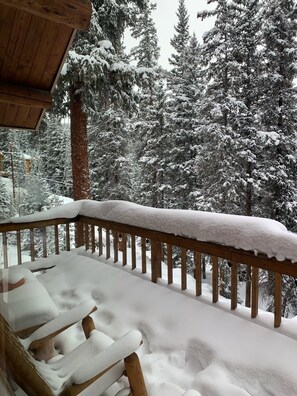 Un balcone in legno con ringhiere imbiancate dalla neve e vista su alberi carichi di neve.