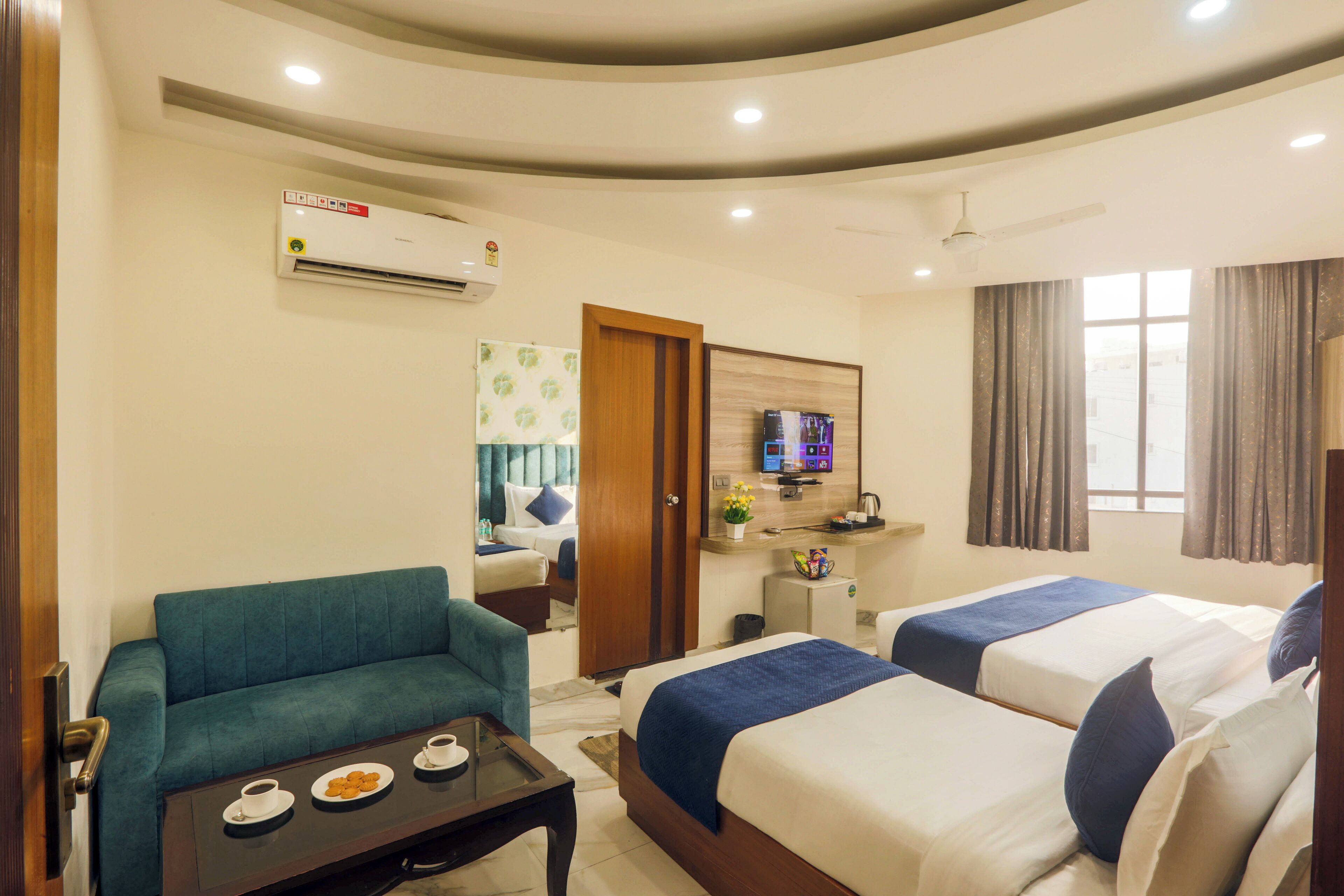 A hotel room with two beds, a sofa, a small table with a cup and cookies, an air conditioner, a TV, and a window with curtains.