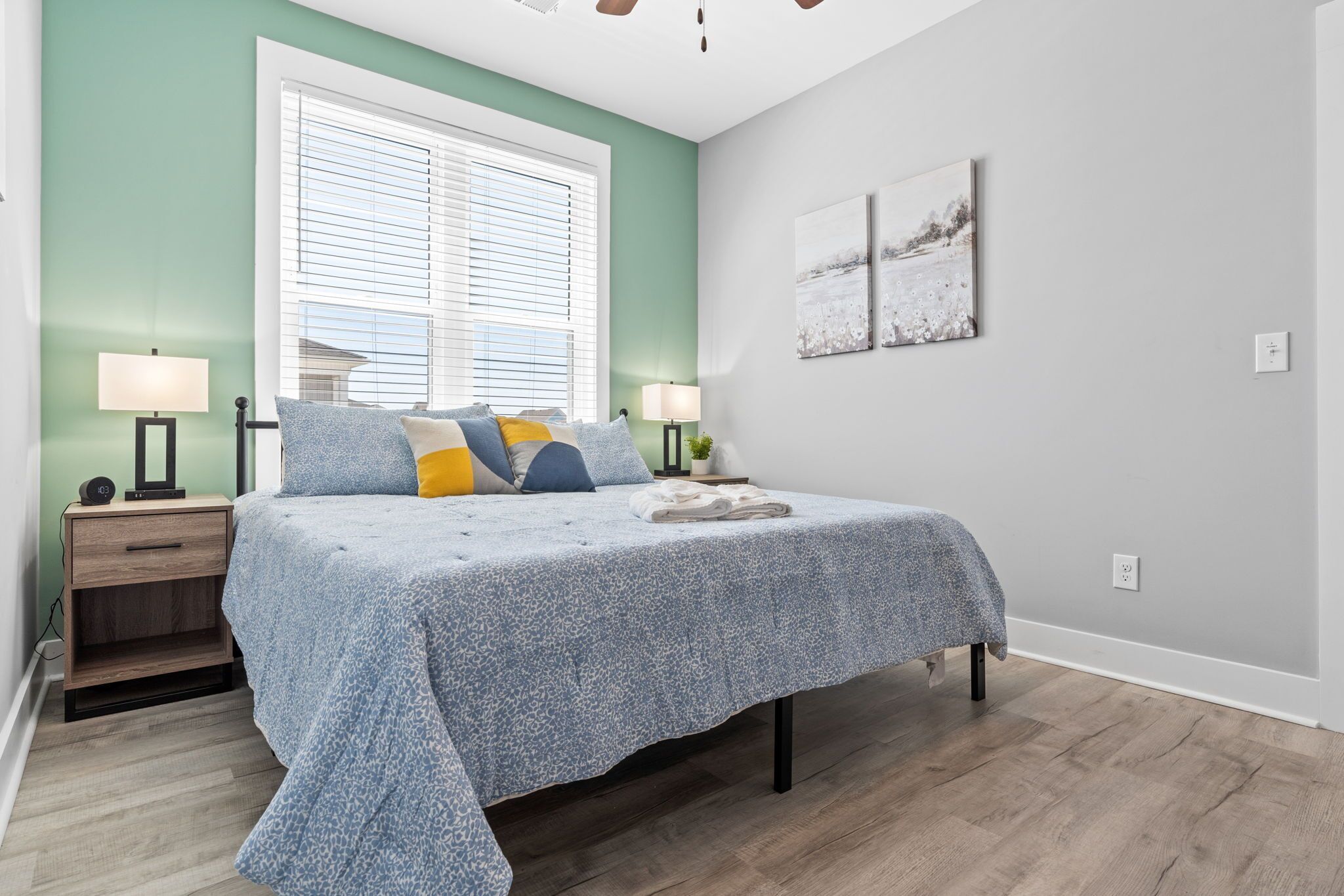 A bedroom with a bed, bedside tables, a window with blinds, and artwork on the wall.