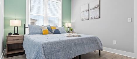 A bedroom with a bed, bedside tables, a window with blinds, and artwork on the wall.