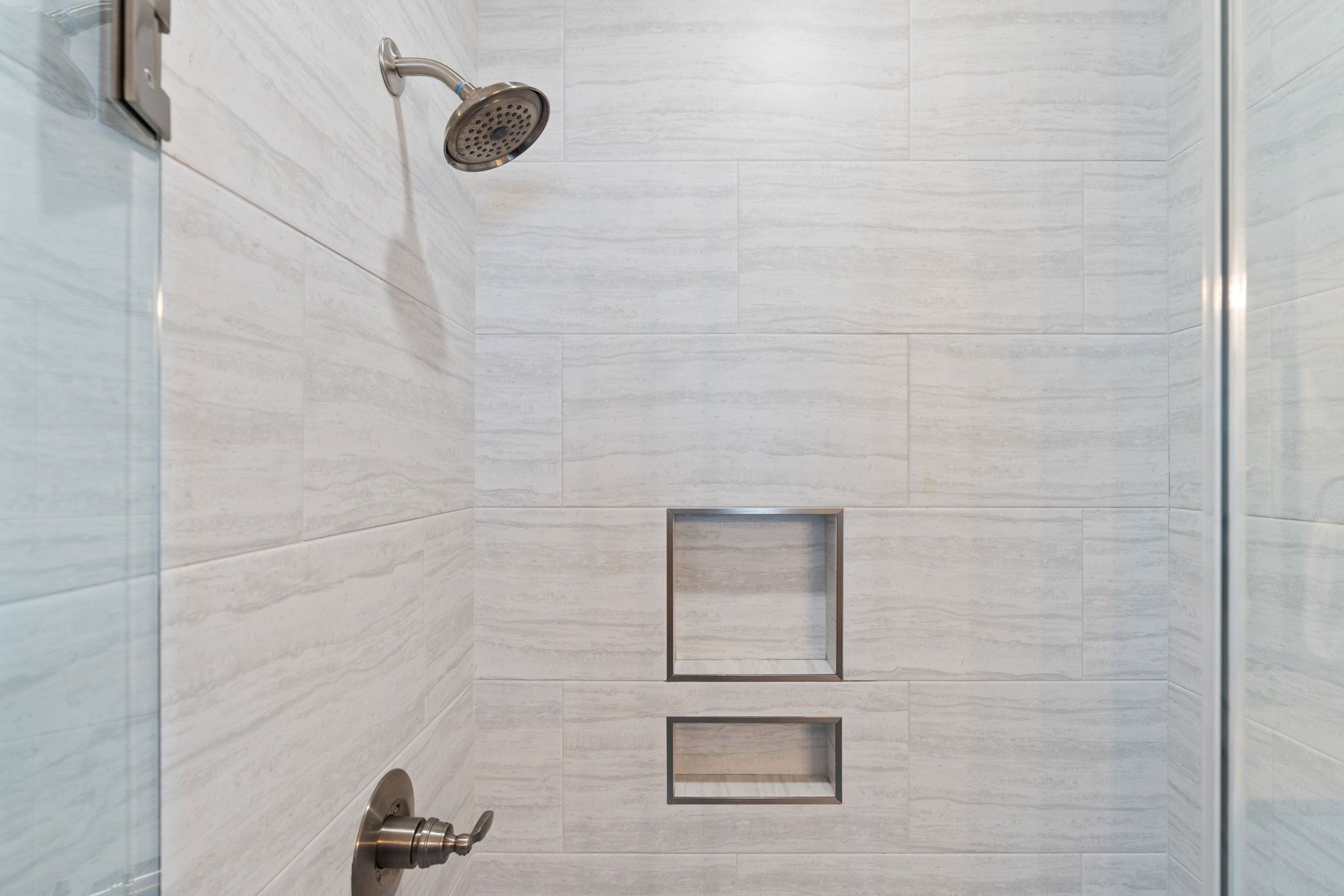 A modern shower with a handheld showerhead, a glass door, and a tiled wall with two rectangular cutouts.