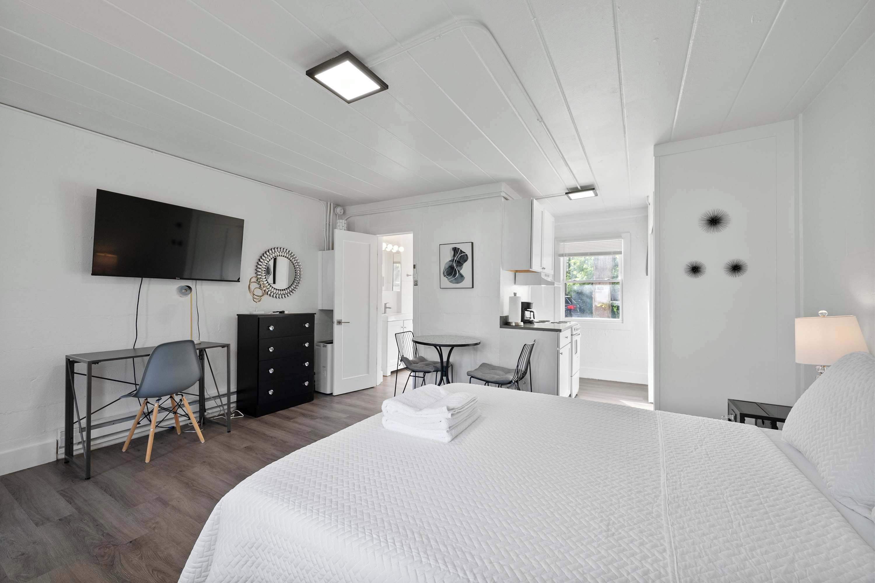 A hotel room with a large bed, a desk with a chair, a television, and a kitchenette area.