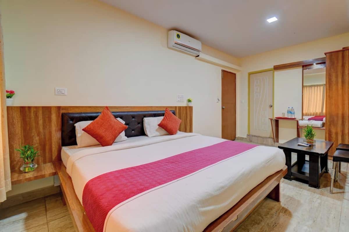 Une chambre d’hôtel avec un grand lit, une tête de lit en bois, une couvre-lit rouge, une petite table avec une plante et un appareil de climatisation fixé au mur.