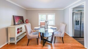 A dining area with a glass table, four chairs, a white cabinet, and a refrigerator in the background.
