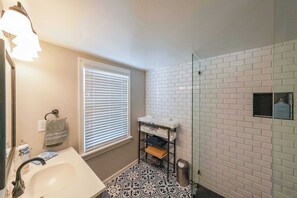 A bathroom with a white sink, a mirror, a towel rack, a window with blinds, a shower area with a glass door, and a patterned floor.