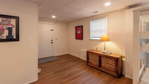 A hallway with wooden flooring, a white door, a framed picture, a wooden cabinet with a lamp, and a window with blinds.