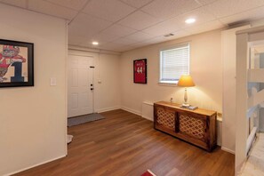 A hallway with wooden flooring, a white door, a framed picture, a wall-mounted sign, a wooden cabinet with a lamp, and a window with blinds.