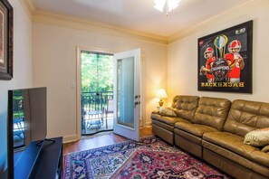 A living room with a leather sofa, a television, a patterned rug, a door leading to a balcony, and a poster on the wall.