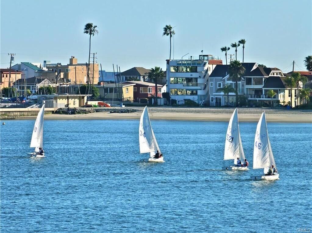A coastal scene with sailboats on calm water, buildings in the background, and a clear sky.