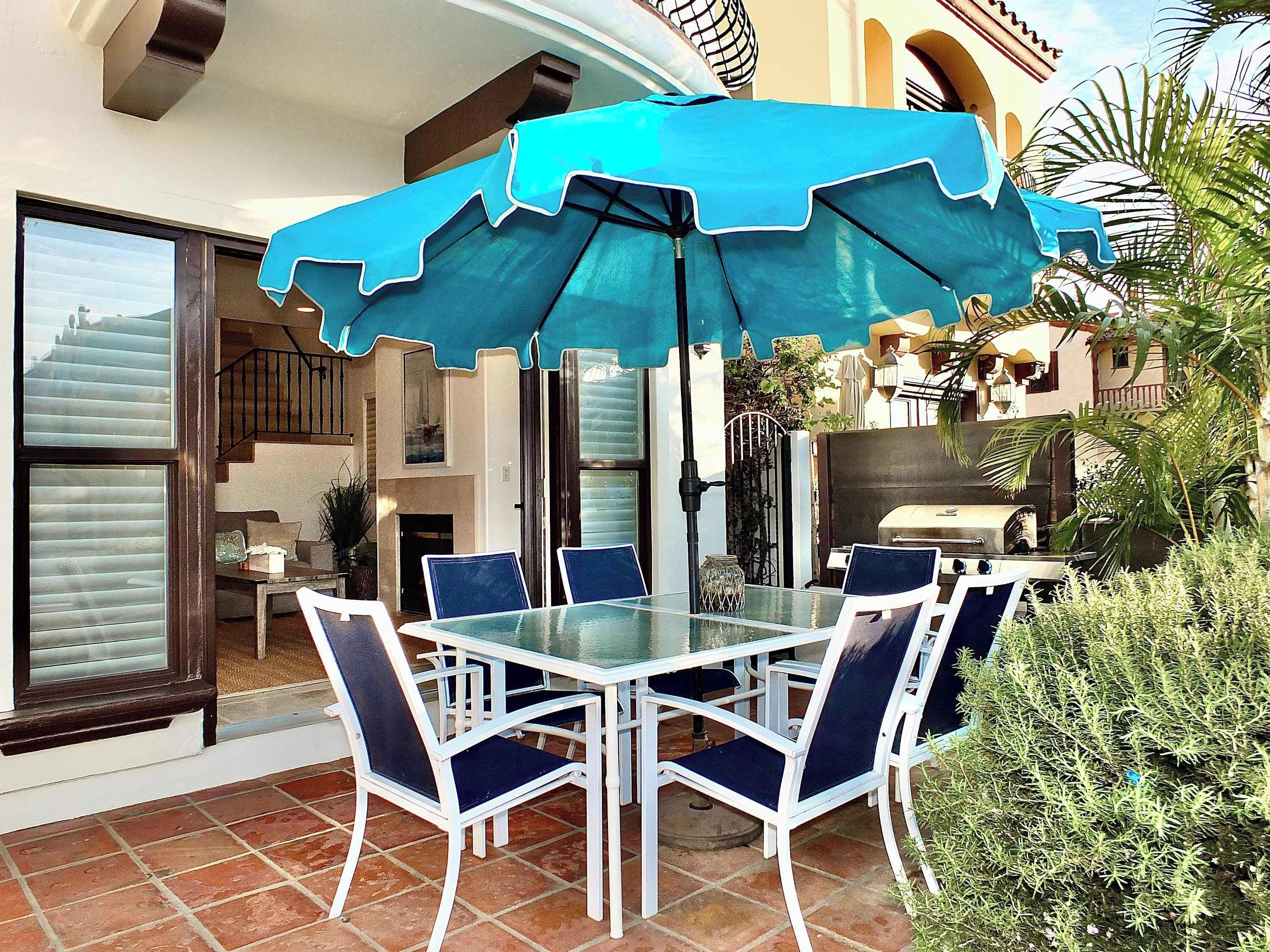 An outdoor patio with a table and chairs, a blue umbrella, and a grill.