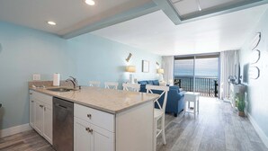 A modern kitchen with a central island, a dining area with white chairs, and a balcony with a view of the ocean.