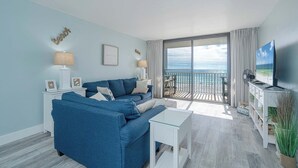 A modern living room with a blue sofa, white side table, and a flat-screen TV. A balcony with a view of the sea is visible.