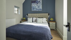 A bedroom with a large bed, a headboard, a nightstand with a lamp, and a wall-mounted painting. - The Hideaway - Sleeps 6 (Bibury)