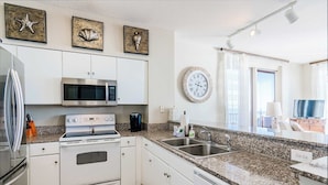 A modern kitchen with white cabinets, a stainless steel refrigerator, a microwave, an oven, and a granite countertop. There are decorative shells on the wall and a clock.