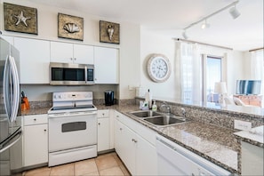 A modern kitchen with white cabinets, a stainless steel refrigerator, a microwave, an oven, and a granite countertop. There are decorative shells on the wall and a clock. - Sunset Shores | Oceanfront, Pool | Activity Experiences (Panama City Beach)
