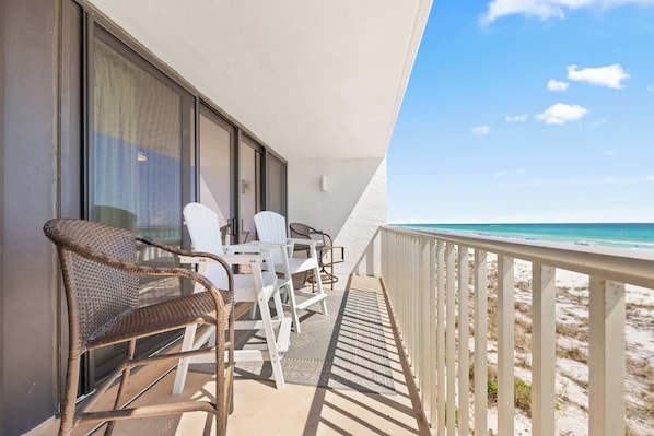 A balcony with a view of the beach, furnished with wicker chairs and white tables.