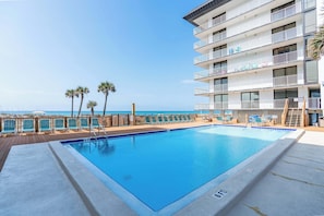 A swimming pool with a view of the ocean, a wooden deck, and a multi-story building with balconies.