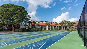 Pistes de tennis i zona de petanca, envoltades per una tanca, amb edificis i palmeres al fons.