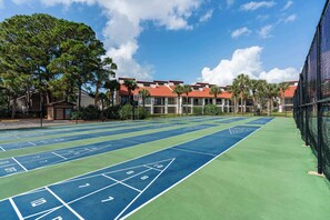 A tennis court with a shuffleboard area, surrounded by a fence, with buildings and palm trees in the background. - Beachfront Days | Ocean View, Beachfront | Activity Experiences (Panama City Beach)