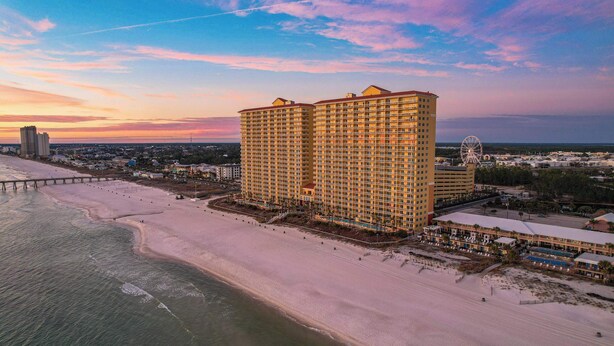A high-rise hotel with multiple floors, a beach in the foreground, and a pier extending into the water. - Sandy Feet Retreat | Ocean View, Beach Access | Activity Deals (Panama City Beach)
