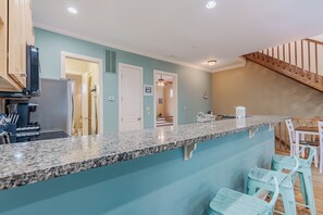 A kitchen with a granite countertop, a refrigerator, and a microwave. There are two light blue bar stools and a wooden staircase in the background. - SeaEsta Luxe Retreat | Pool, Walk to Beach | Activity Opportunities (Panama City Beach)
