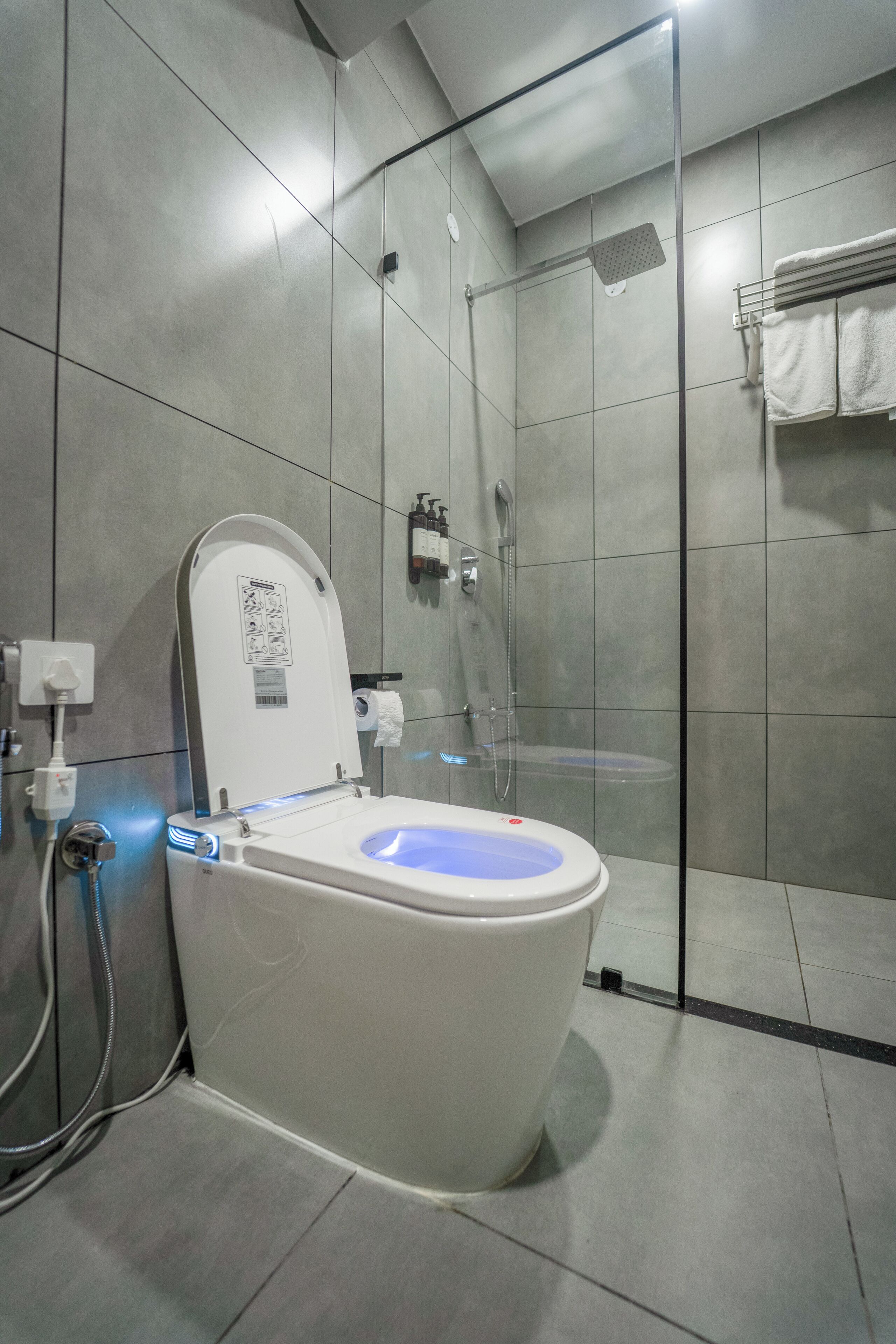 Een moderne badkamer met een douchecabine van glas, een wit toilet met een blauw verlicht zitvlak, en grijze betegelde wanden.