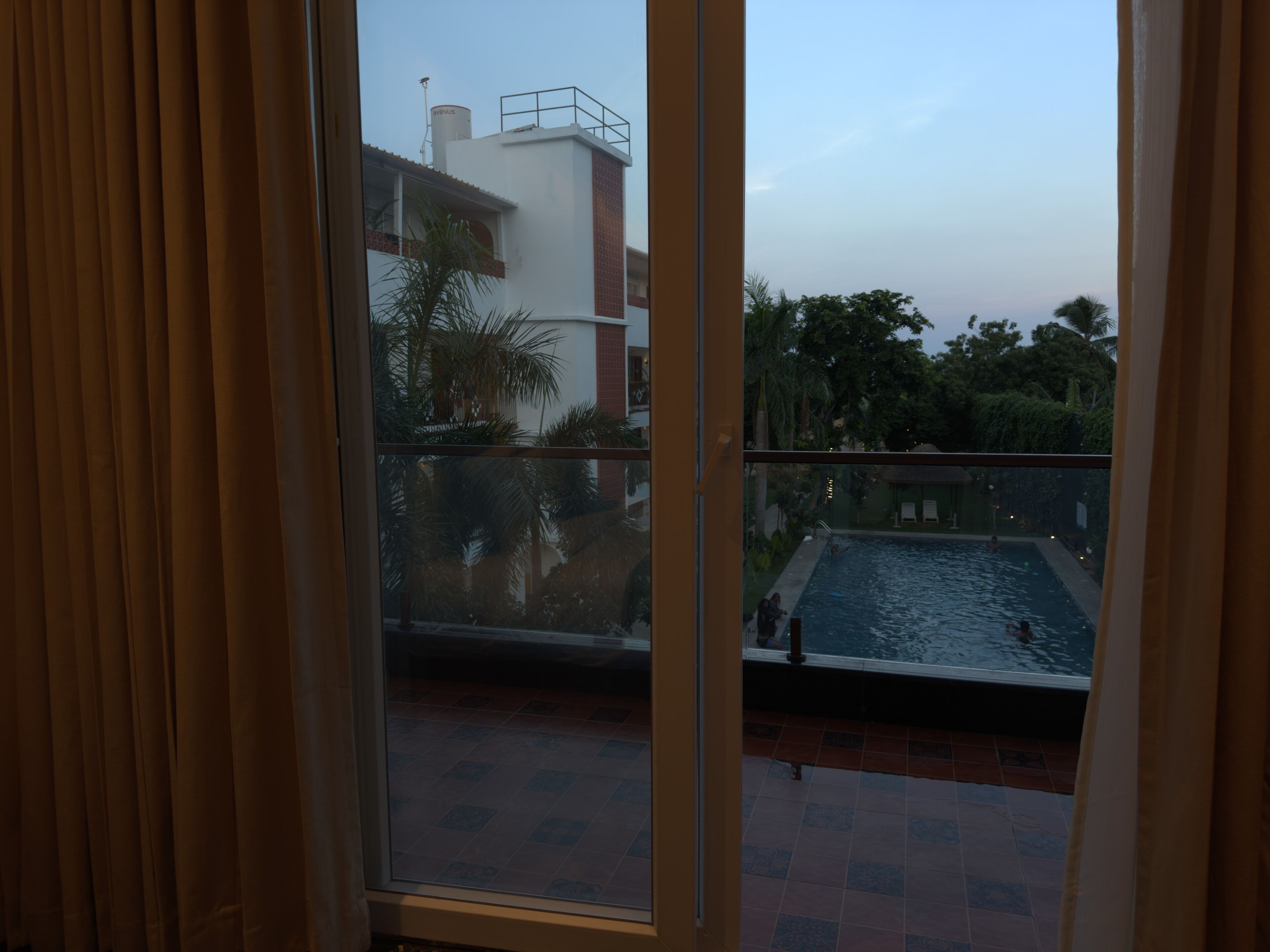 A view through a window of a residential building with a swimming pool, a tiled floor, and a balcony with a railing.