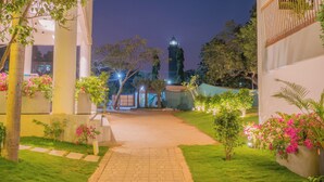 Vista nocturna de un patio con un sendero central, rodeado de vegetación y un edificio con estructura de cúpula.