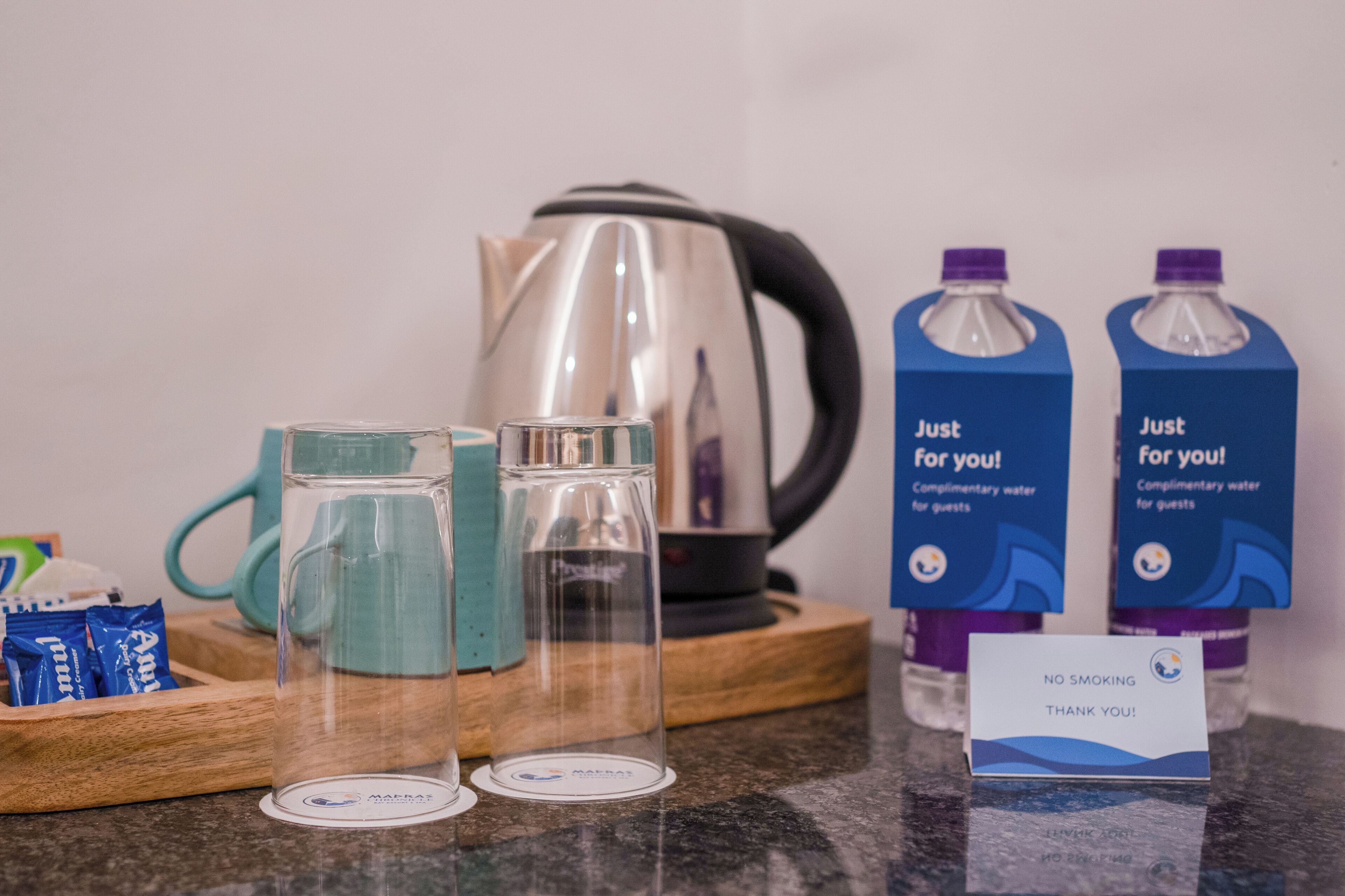 A hotel room's breakfast setup with a kettle, two glass mugs, two water bottles, a tray with packets, and a 'No Smoking' sign.