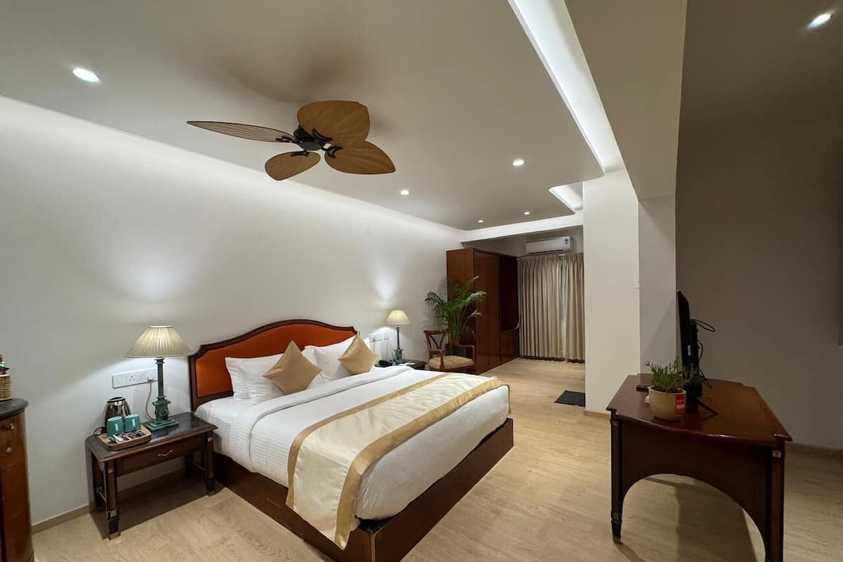 Una habitación de hotel moderna con una cama grande, cabecera de madera, mesitas de noche con lámparas, ventilador de techo y un escritorio con una planta.